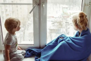 Two children sitting near a window. One of them is wrapped in a blue blanket.